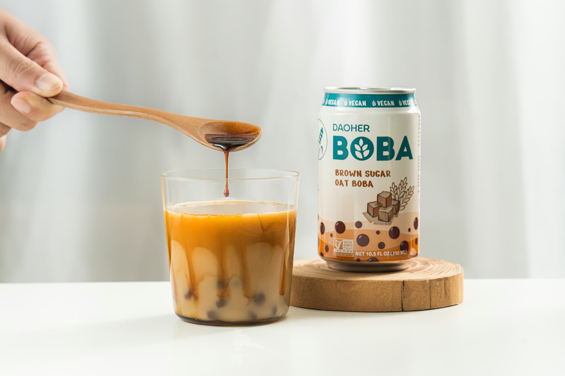DaoHer Brown Sugar Oat BOBA beside a glass of milk tea as brown sugar syrup drips from a wooden spoon